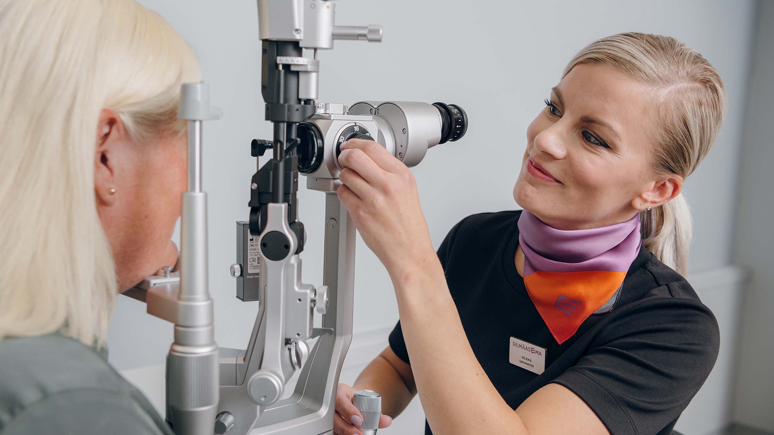 An optician adjusts a microscope in an eye examination room while the customer sits opposite.